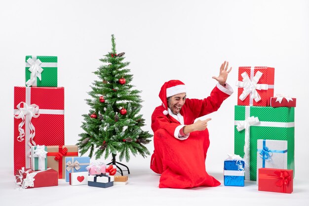 Jovem engraçado sorridente vestido de Papai Noel com presentes e uma árvore de Natal decorada sentado no chão apontando algo no lado esquerdo sobre fundo branco