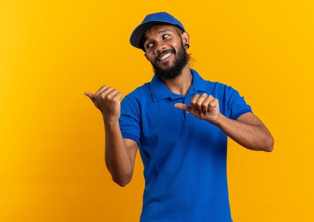 Jovem entregador afro-americano sorridente, olhando e apontando para o lado isolado em um fundo laranja com espaço de cópia