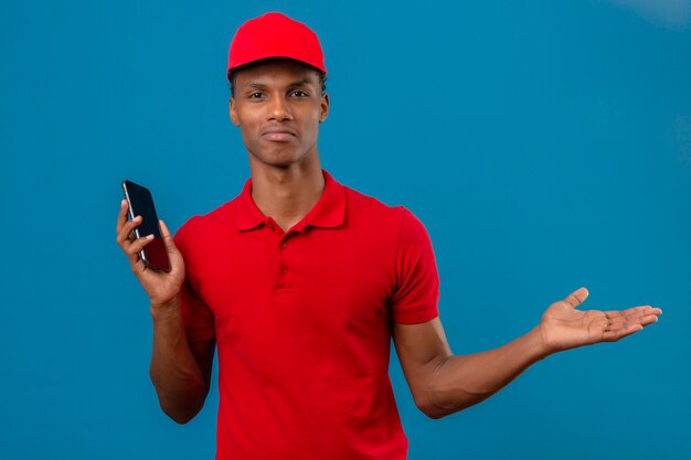 Jovem entregador americano africano vestindo camisa polo vermelha e boné em pé com o smartphone na mão, olhando confuso e tendo dúvidas sobre azul isolado