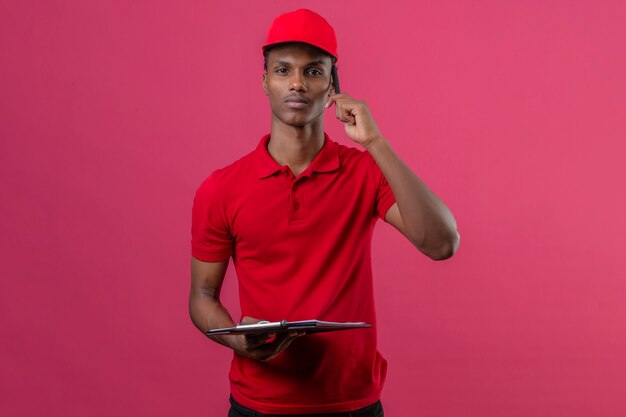 Jovem entregador americano africano vestindo camisa polo vermelha e boné segurando a área de transferência e specking pelo smartphone com cara séria sobre rosa isolado