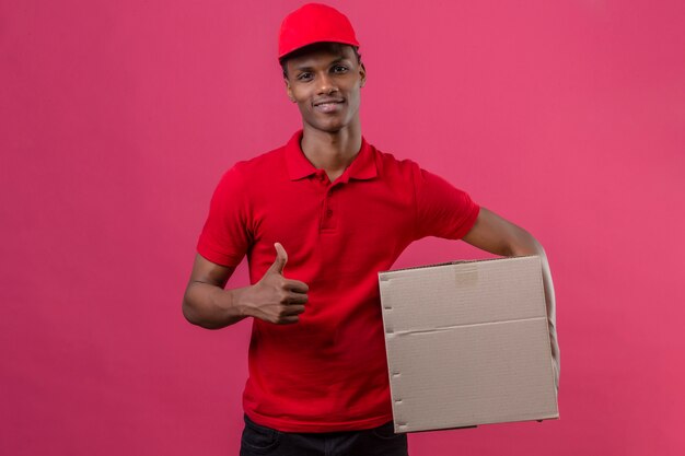 Jovem entregador americano africano vestindo camisa polo vermelha e boné segurando a caixa de papelão aparecendo polegar com sorriso no rosto sobre rosa isolado