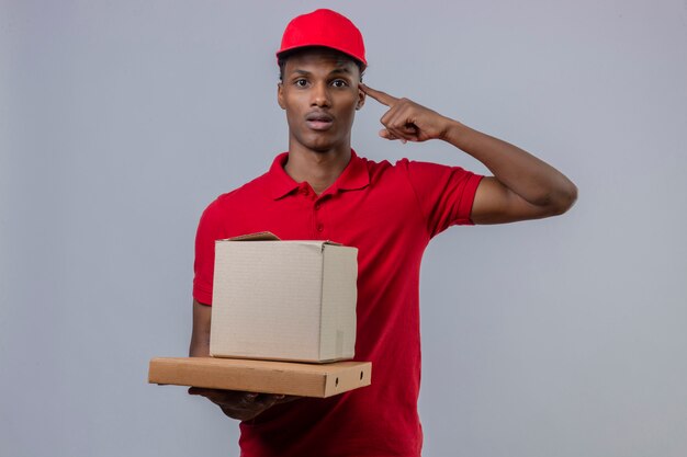 Jovem entregador americano africano vestindo camisa polo vermelha e boné segurando a pilha de caixas de papelão, apontando para a cabeça com um dedo sobre branco isolado