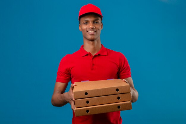 Jovem entregador americano africano vestindo camisa polo vermelha e boné segurando a pilha de caixas de pizza com um grande sorriso sobre azul isolado