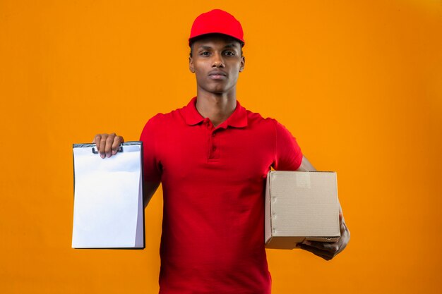 Jovem entregador americano africano vestindo camisa polo vermelha e boné segurando o pacote caixa e área de transferência com espaços em branco vazios, olhando confiante sobre laranja isolado