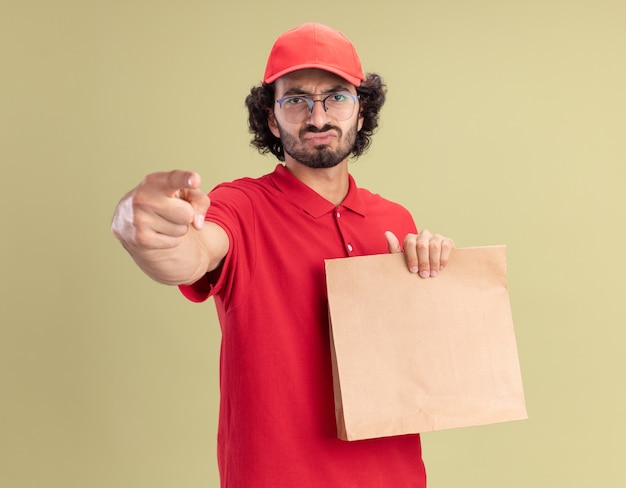 Jovem entregador caucasiano, carrancudo, de uniforme vermelho e boné de óculos, segurando um pacote de papel, olhando e apontando isolado na parede verde oliva