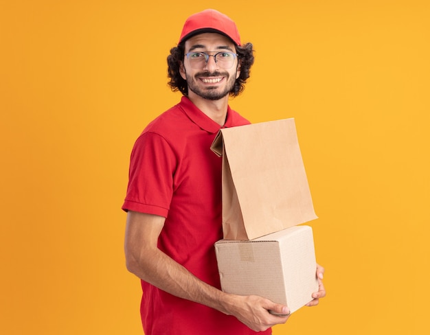jovem entregador caucasiano, sorridente, de uniforme vermelho e boné, usando óculos, de pé na vista de perfil, segurando uma caixa de papelão com um pacote de papel olhando para a frente