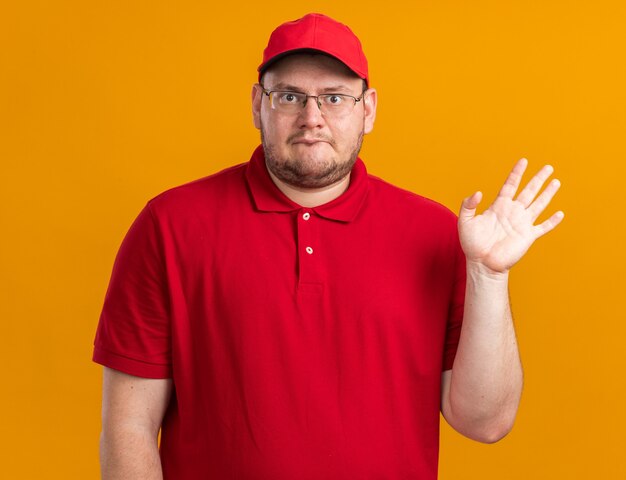 jovem entregador com excesso de peso impressionado com óculos ópticos em pé com a mão levantada, isolado em uma parede laranja com espaço de cópia