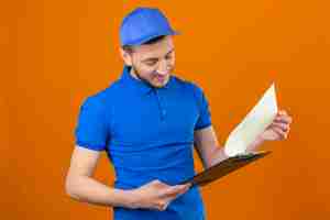 Foto grátis jovem entregador de camisa pólo azul e boné olhando para a prancheta nas mãos com cara séria sobre fundo laranja isolado
