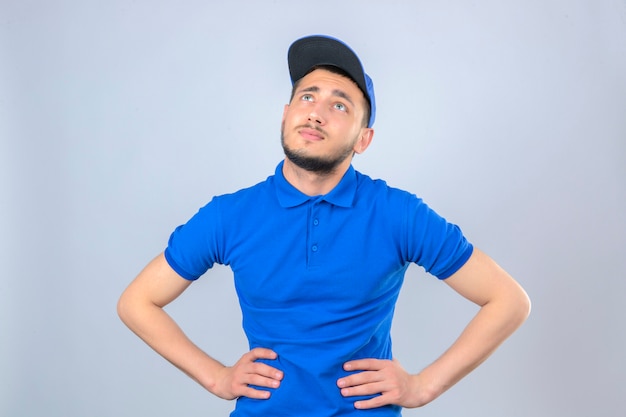 Foto grátis jovem entregador de camisa pólo azul e boné parecendo entediado olhando para cima sobre um fundo branco isolado