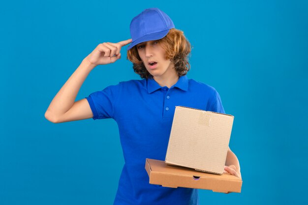 Jovem entregador de camisa pólo azul e boné segurando caixas de papelão apontando para a cabeça com um dedo uma ótima ideia ou pensamento em pé sobre um fundo azul isolado