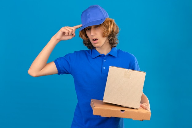 Jovem entregador de camisa pólo azul e boné segurando caixas de papelão apontando para a cabeça com um dedo uma ótima ideia ou pensamento em pé sobre um fundo azul isolado