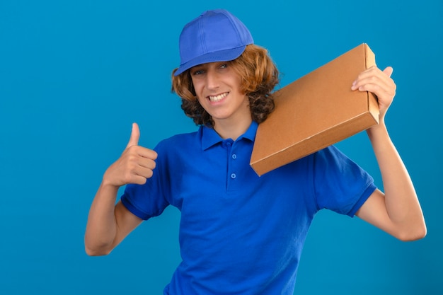 Jovem entregador de camisa pólo azul e boné segurando uma caixa de pizza no ombro e mostrando o polegar sorrindo alegremente em pé sobre um fundo azul isolado