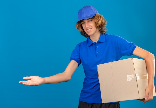 Jovem entregador de camisa pólo azul e boné segurando uma grande caixa de papelão apontando com a palma da mão, olhando para a câmera sobre fundo azul isolado