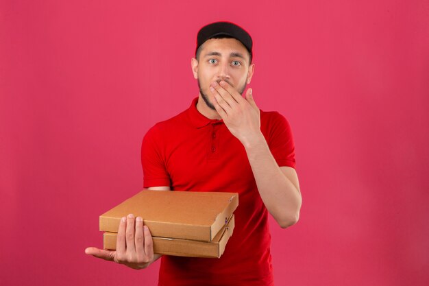 Jovem entregador de camisa pólo vermelha e boné em pé com caixas de pizza, parecendo surpreso e chocado, cobrindo a boca sobre um fundo rosa isolado