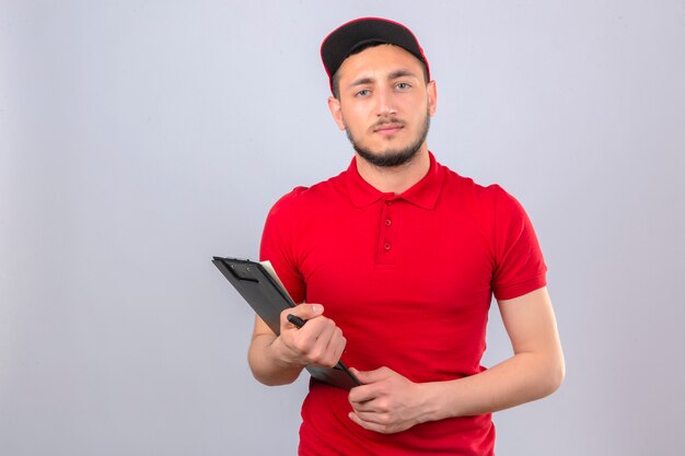 Jovem entregador de camisa pólo vermelha e boné em pé com uma pilha de caixas de pizza apontando para elas com uma cara séria sobre um fundo branco isolado