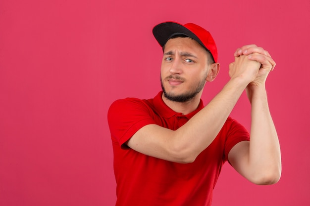 Foto grátis jovem entregador de camisa pólo vermelha e boné gesticulando com as mãos fechadas, parecendo confiante e orgulhoso sobre um fundo rosa isolado