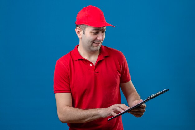 Jovem entregador de camisa pólo vermelha e boné olhando para a prancheta nas mãos com um sorriso no rosto em pé sobre um fundo azul isolado