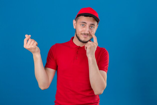 Jovem entregador de camisa pólo vermelha e boné preocupado com dinheiro, fazendo um gesto de dinheiro com a mão sobre fundo azul isolado