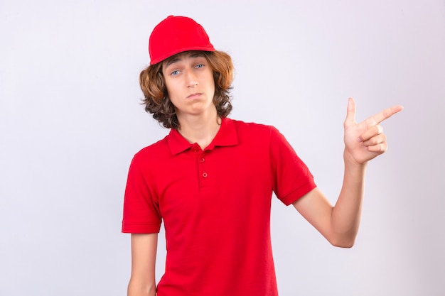 Jovem entregador de uniforme vermelho decepcionado apontando com a mão e o dedo para o lado sobre um fundo branco isolado