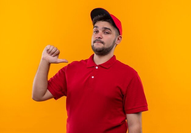 Jovem entregador de uniforme vermelho e boné apontando para si mesmo satisfeito