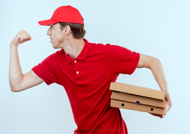 Foto grátis jovem entregador de uniforme vermelho e boné correndo para entregar caixas de pizza para um cliente na parede branca