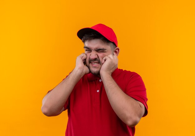 Jovem entregador de uniforme vermelho e boné fechando eras com dedos com expressão irritada pelo barulho