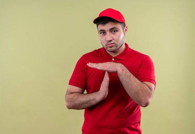 Jovem entregador de uniforme vermelho e boné parecendo cansado fazendo gesto de pausa com as mãos em pé sobre a parede verde