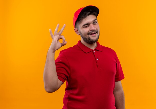 Jovem entregador de uniforme vermelho e boné piscando e sorrindo mostrando sinal de ok