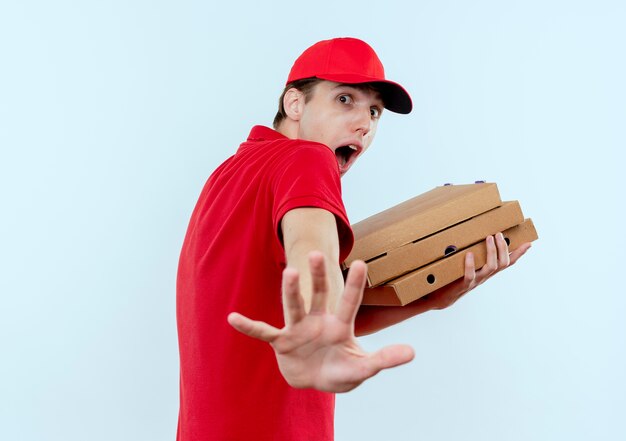 Jovem entregador de uniforme vermelho e boné segurando caixas de pizza com muito medo, fazendo gesto de defesa com a mão em pé sobre uma parede branca