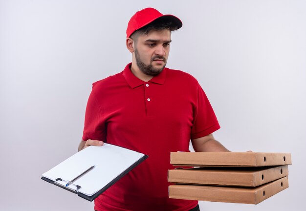 Jovem entregador de uniforme vermelho e boné segurando caixas de pizza e prancheta com caneta e páginas em branco, parecendo confuso tentando fazer uma escolha