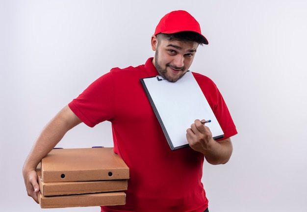 Jovem entregador de uniforme vermelho e boné segurando caixas de pizza mostrando a prancheta com páginas em branco pedindo assinatura