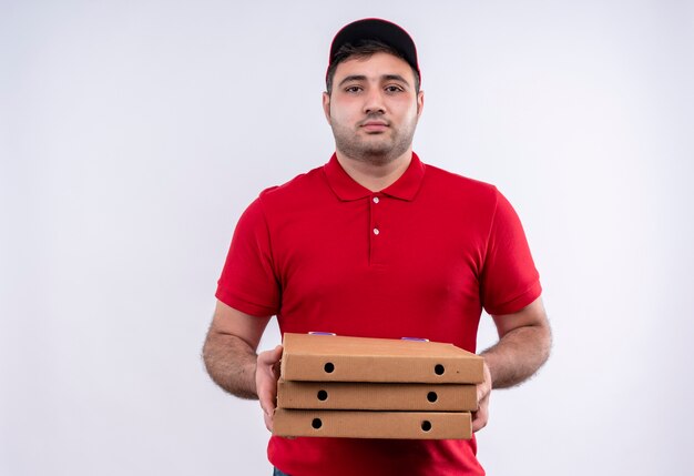 Jovem entregador de uniforme vermelho e boné segurando caixas de pizza, olhando para a câmera com expressão confiante em pé sobre uma parede branca