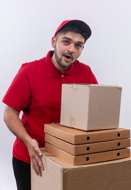 Jovem entregador de uniforme vermelho e boné segurando grandes caixas de papelão parecendo surpreso e surpreso