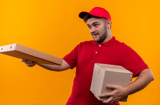 Jovem entregador de uniforme vermelho e boné segurando um pacote de caixa dando uma caixa de pizza para um cliente