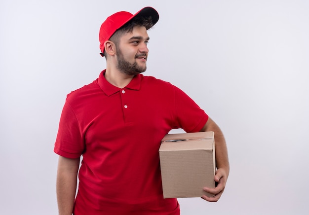 Jovem entregador de uniforme vermelho e boné segurando um pacote de caixa olhando para o lado e sorrindo confiante