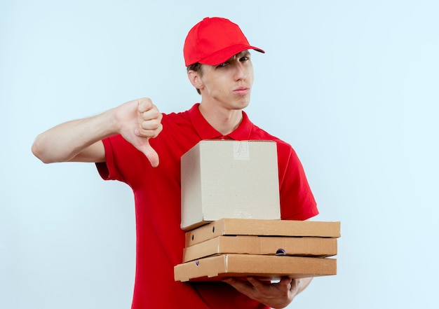 Jovem entregador de uniforme vermelho e boné segurando um pacote de caixas e caixas de pizza olhando para a frente descontente mostrando polegares para baixo em pé sobre uma parede branca