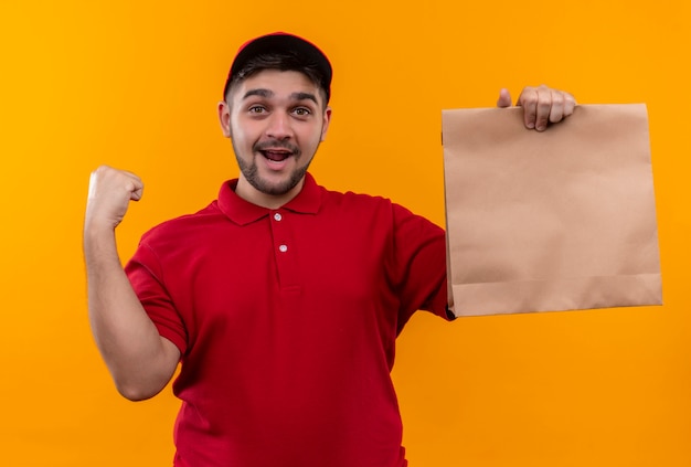 Jovem entregador de uniforme vermelho e boné segurando um pacote de papel sorrindo alegremente levantando o punho feliz e animado
