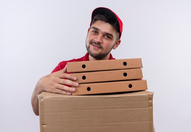 Jovem entregador de uniforme vermelho e boné segurando uma pilha de caixas de pizza e uma caixa de papelão, sorrindo amigável, olhando para a câmera