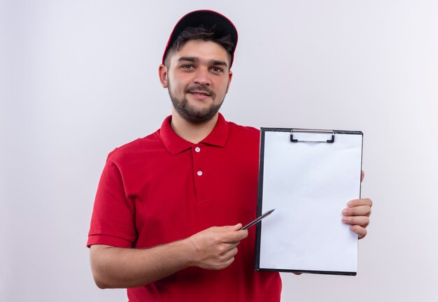 Jovem entregador de uniforme vermelho e boné segurando uma prancheta com páginas em branco apontando com uma caneta para ele, pedindo assinatura