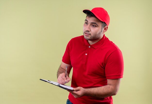 Jovem entregador de uniforme vermelho e boné segurando uma prancheta escrevendo algo olhando para o lado com uma cara séria em pé sobre a parede verde