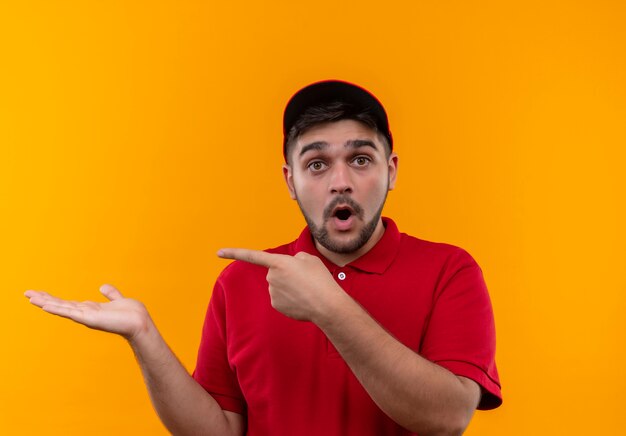 Jovem entregador de uniforme vermelho e boné surpreso apontando com o braço e o dedo para o lado