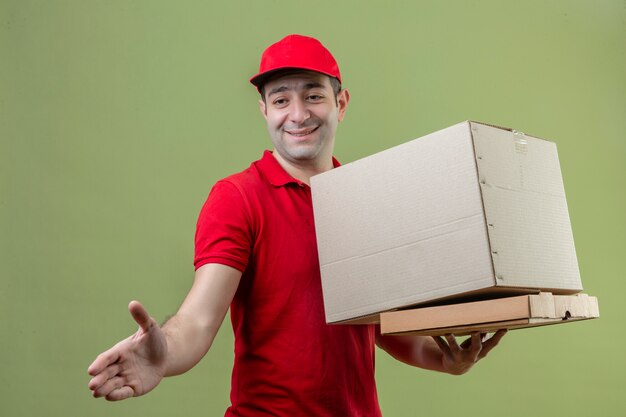 Jovem entregador de uniforme vermelho, fazendo gesto de saudação, oferecendo a mão, segurando caixas de papelão e sorrindo sobre fundo verde isolado