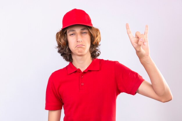 Jovem entregador de uniforme vermelho fazendo o símbolo do rock com as mãos ao alto orgulhoso e confiante sobre fundo branco isolado