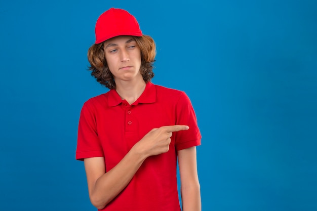 Jovem entregador de uniforme vermelho olhando para o lado apontando o dedo para algo ao lado cético e nervoso sobre fundo azul isolado