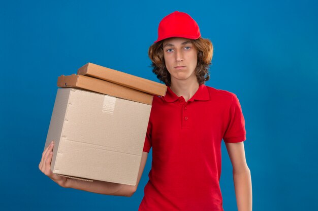 Jovem entregador de uniforme vermelho segurando caixas de papelão, olhando para a câmera, nervoso e cético em pé sobre um fundo azul isolado