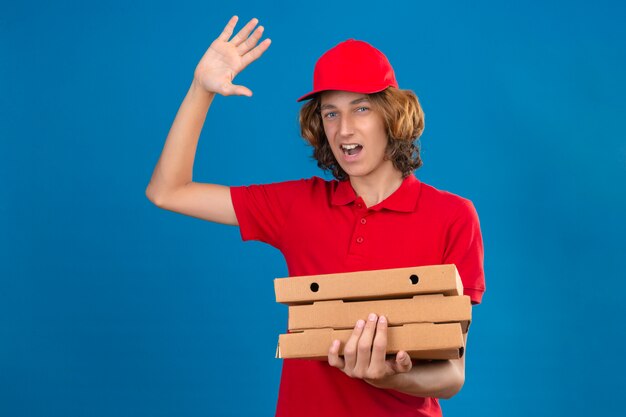 Jovem entregador de uniforme vermelho segurando caixas de pizza acenando com a mão e sorrindo alegremente sobre um fundo azul isolado