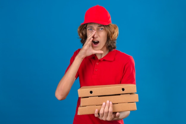 Jovem entregador de uniforme vermelho segurando caixas de pizza segurando a mão perto da boca aberta gritando sobre um fundo azul isolado