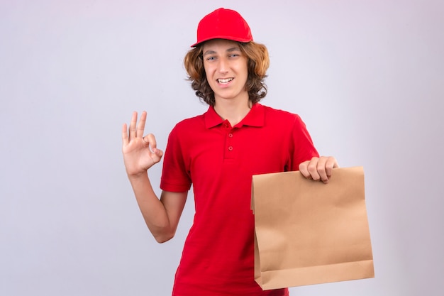 Jovem entregador de uniforme vermelho segurando um pacote de papel, fazendo sinal de ok, sorrindo alegremente sobre fundo branco isolado