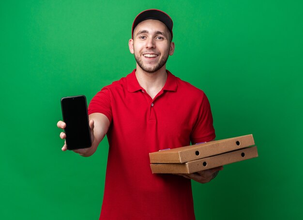 Jovem entregador sorridente, usando uniforme com tampa, segurando caixas de pizza com telefone