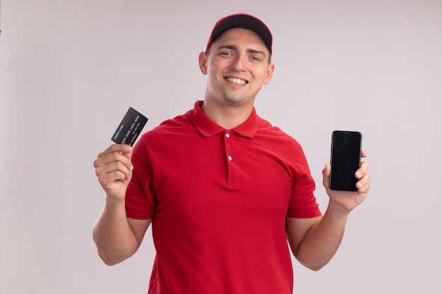 Jovem entregador sorridente, vestindo uniforme com tampa, segurando o cartão de crédito com telefone isolado na parede branca